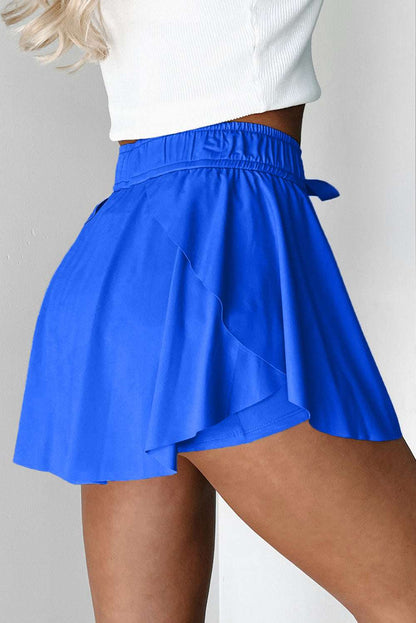 Blue fabric tennis skort with an elastic waistband, drawstring tie, and layered ruffle hem shown from a side view.