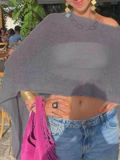 A woman outdoors wearing a gray knit poncho-style top with an asymmetrical one-shoulder design, paired with blue jeans and holding a bright pink fringed bag.