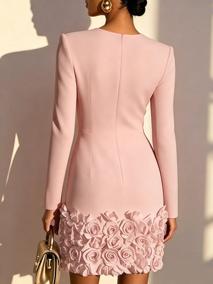 A woman shown from the back wearing a fitted light pink long-sleeve dress with a rose-patterned hem, holding a small metallic handbag against a sunlit neutral wall.