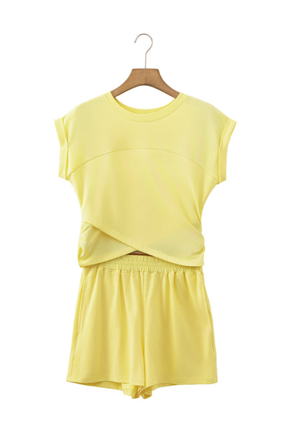 A two-piece women's lounge set in pale yellow, featuring a short-sleeve crewneck top with a crossover front detail and matching shorts, displayed on a wooden hanger against a white background.