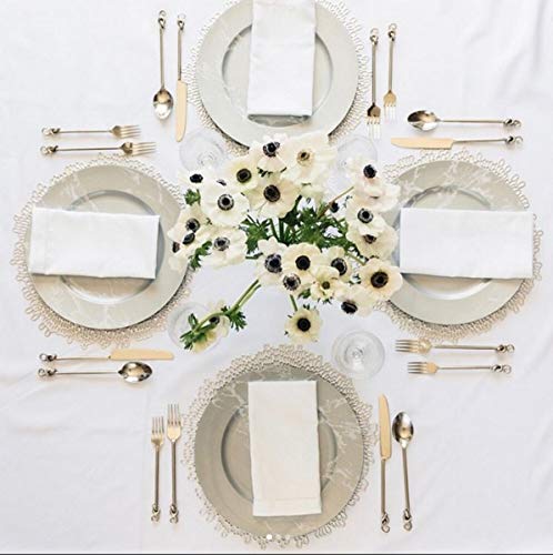 An overhead flat-lay of a formal table setting on a white tablecloth, featuring four white plates with scalloped clear lace rims, white folded napkins, silver flatware, and a central bouquet of white anemone flowers.