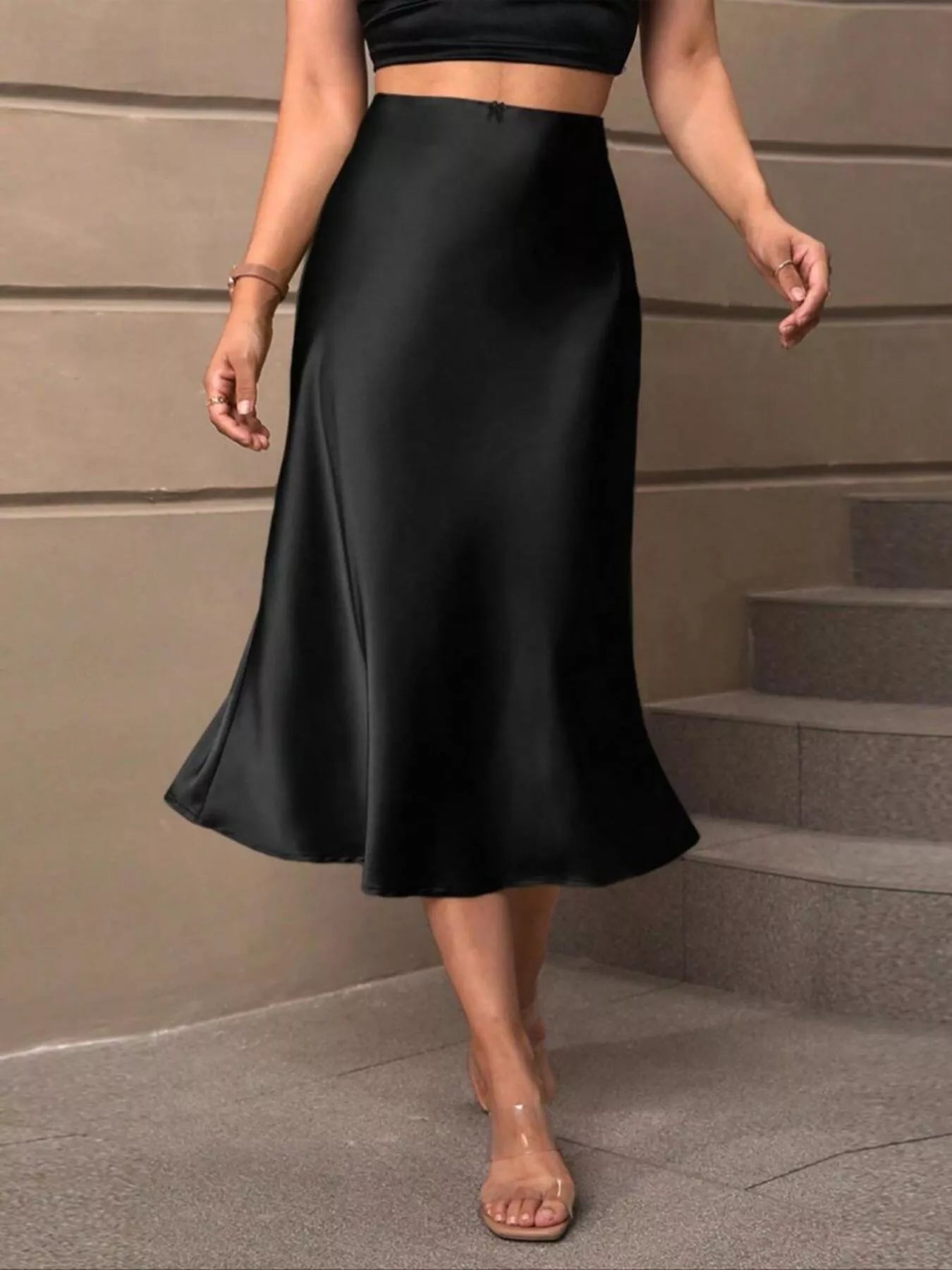 A woman wearing a black satin midi skirt with a flared hem, paired with a black crop top and clear high-heel sandals, standing on stone steps.
