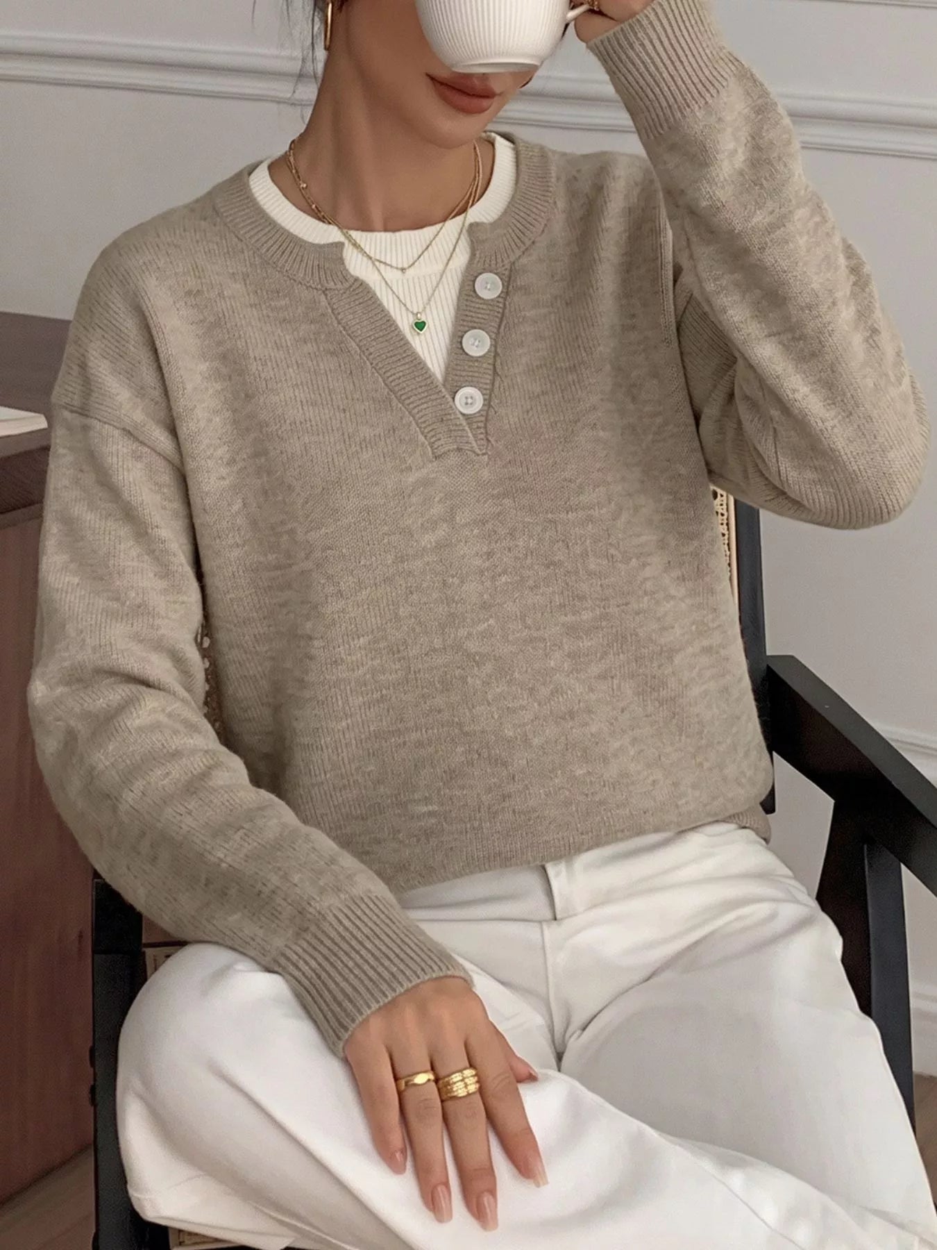 A woman wearing a light taupe knit henley sweater over a white crewneck top and white pants, sitting in a chair and holding a white mug.