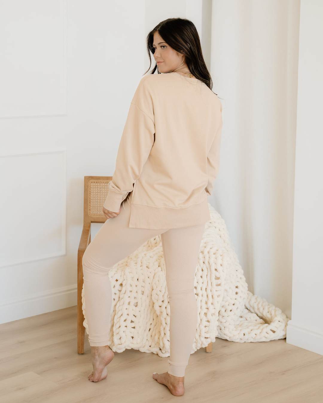 Woman in a beige outfit with a white textured garment, standing in a minimalistic room.