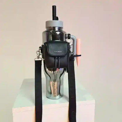 COLDEST water bottle in silver with a black mini backpack holder and pink pen attached, displayed on a white pedestal.