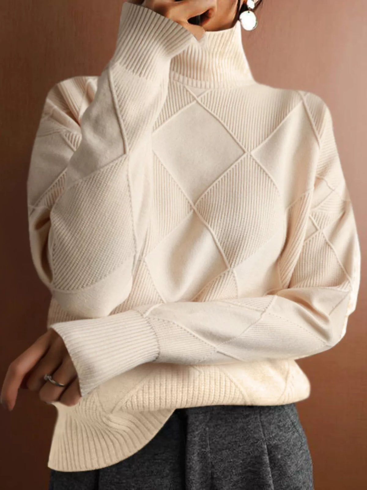 A close-up of a person wearing a cream-colored knit turtleneck sweater with a diamond argyle pattern, paired with dark gray pants, against a warm brown background.