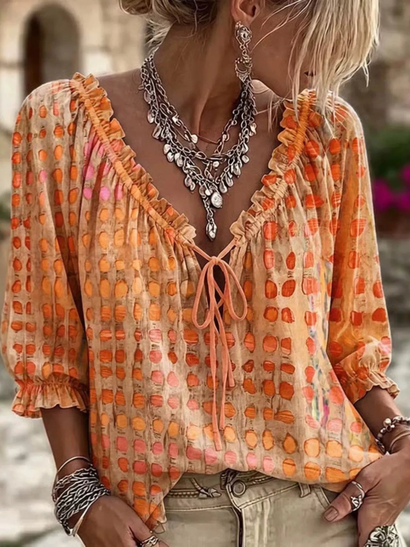 A woman wearing a loose, long-sleeve tie-front blouse with an orange and peach abstract dot pattern on a beige base, styled with layered silver necklaces and bracelets.