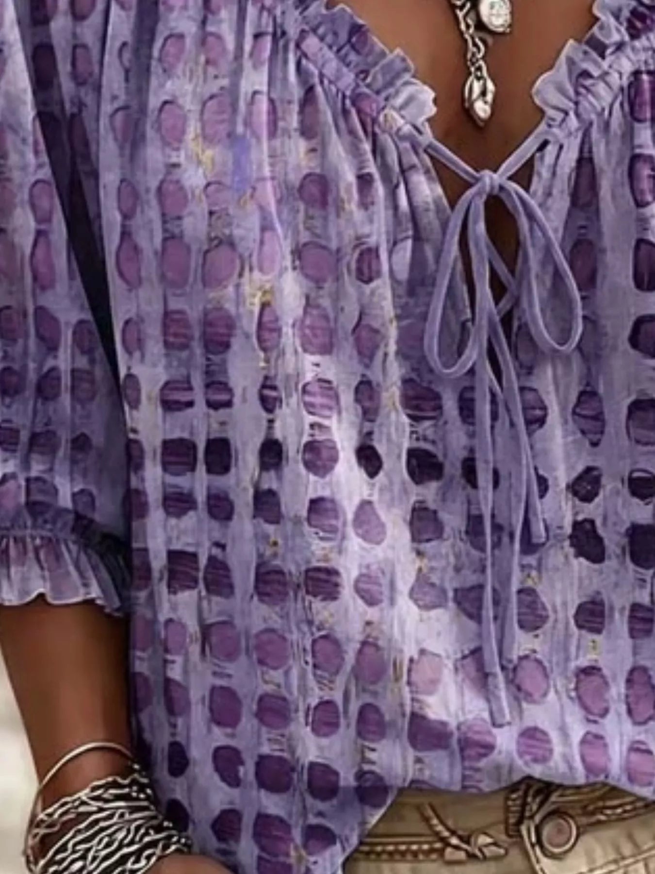 A close-up of a person wearing a lavender and purple patterned blouse with a ruffled neckline, tie-front closure, and elbow-length sleeves. The blouse has an abstract dotted or watercolor-like print in shades of purple and lavender.