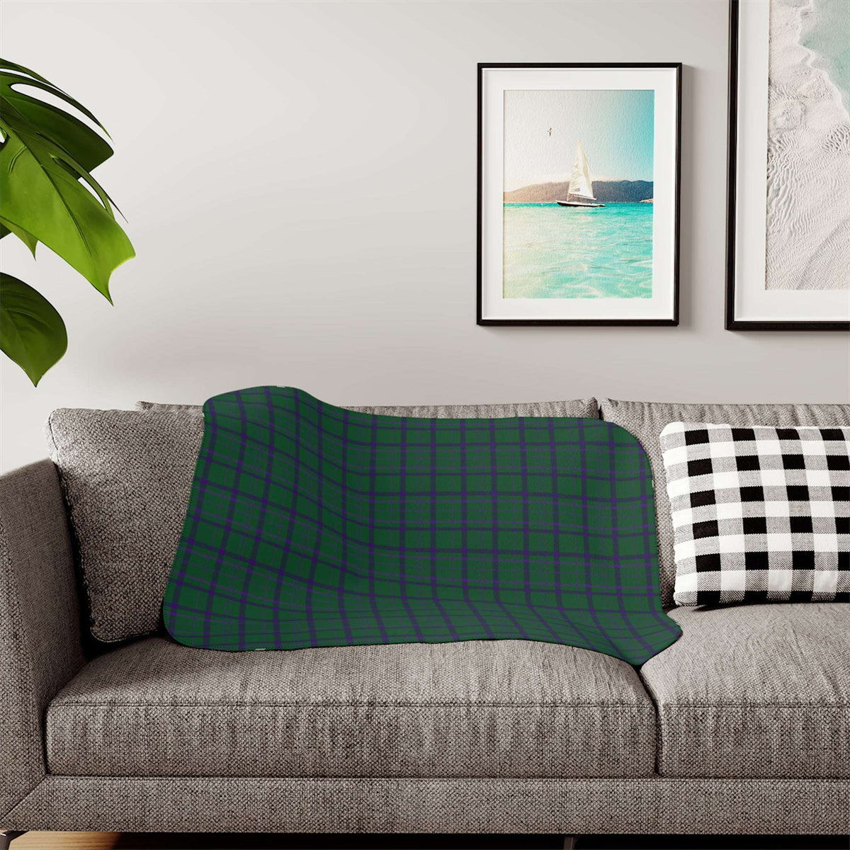 A patterned throw blanket draped over a gray fabric sofa in a living room setting, featuring a botanical design in deep teal, navy, and cream. A black and white checkered pillow sits on the right, with framed wall art and a green plant leaf visible in the background.