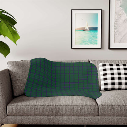 A patterned throw blanket draped over a gray fabric sofa in a living room setting, featuring a botanical design in deep teal, navy, and cream. A black and white checkered pillow sits on the right, with framed wall art and a green plant leaf visible in the background.