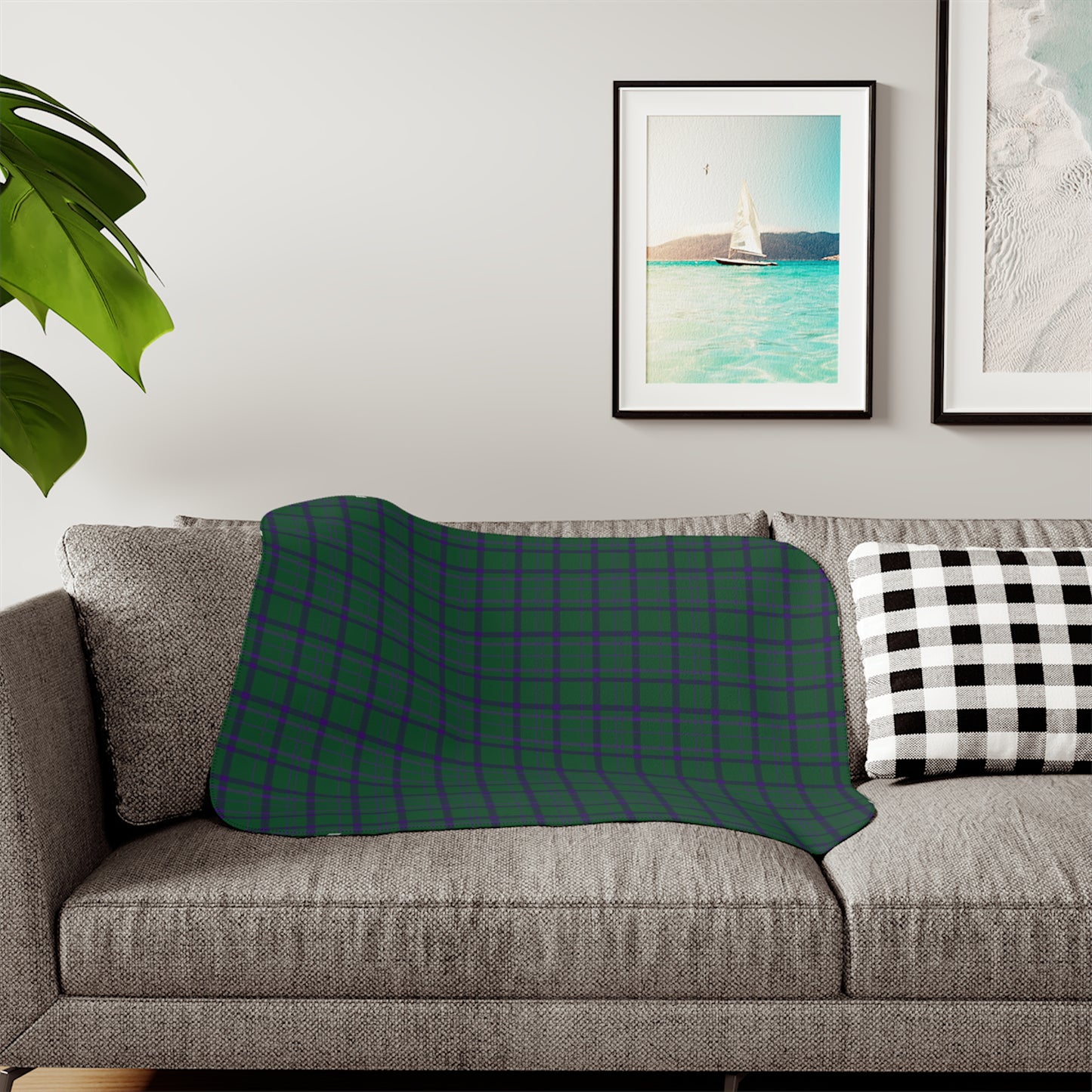 A patterned throw blanket draped over a gray fabric sofa in a living room setting, featuring a botanical design in deep teal, navy, and cream. A black and white checkered pillow sits on the right, with framed wall art and a green plant leaf visible in the background.
