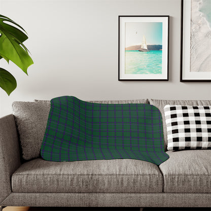 A patterned throw blanket draped over a gray fabric sofa in a living room setting, featuring a botanical design in deep teal, navy, and cream. A black and white checkered pillow sits on the right, with framed wall art and a green plant leaf visible in the background.