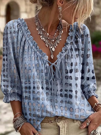 A woman wearing a light blue bohemian-style blouse with a dark gray abstract pattern, ruffled neckline, and tie-front detail, styled with layered silver necklaces and bracelets.