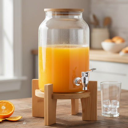 Clear glass beverage dispenser with orange liquid, a metal spigot, and a wood lid and stand on a table.