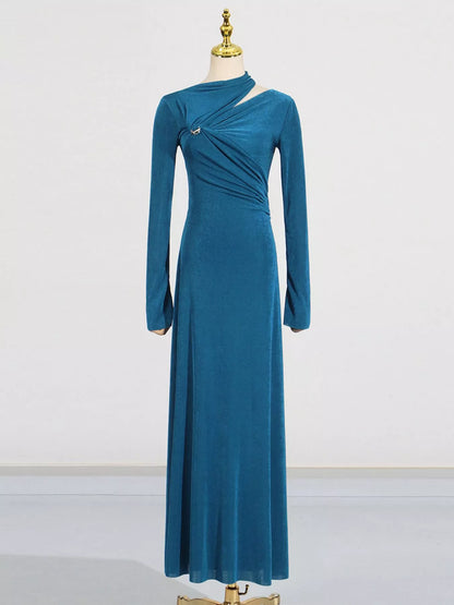 A long-sleeve teal blue velvet maxi dress with an asymmetrical draped neckline and a small silver brooch, displayed on a gold mannequin against a white studio background.