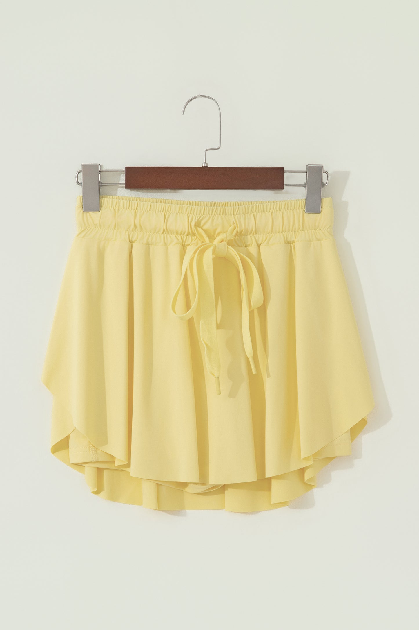 A pair of light yellow high-low shorts with an elastic waistband and a matching drawstring tie, hanging on a wooden hanger against a plain off-white wall.