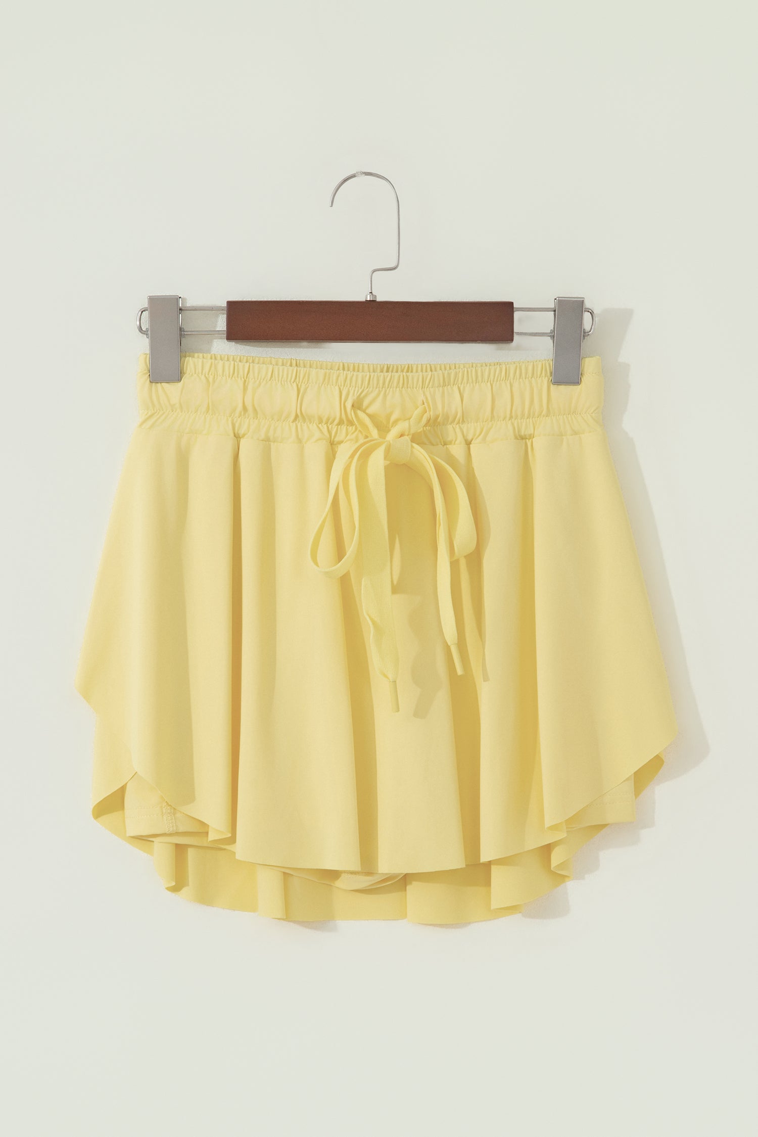A pair of light yellow high-low shorts with an elastic waistband and a matching drawstring tie, hanging on a wooden hanger against a plain off-white wall.