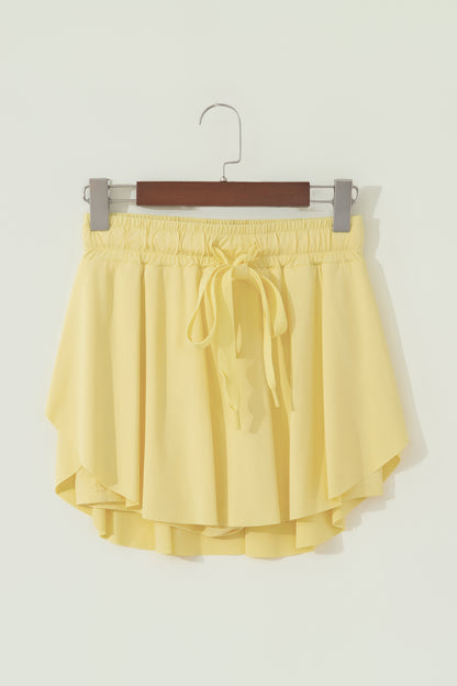 A pair of light yellow high-low shorts with an elastic waistband and a matching drawstring tie, hanging on a wooden hanger against a plain off-white wall.