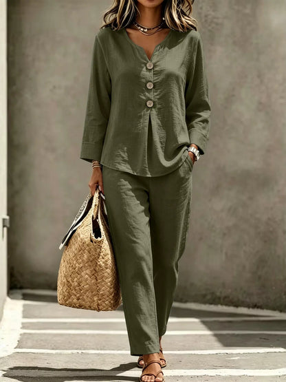 A woman wearing an olive green two-piece linen set with a long-sleeve button-front top and matching wide-leg pants, holding a woven straw tote bag against a textured gray wall.