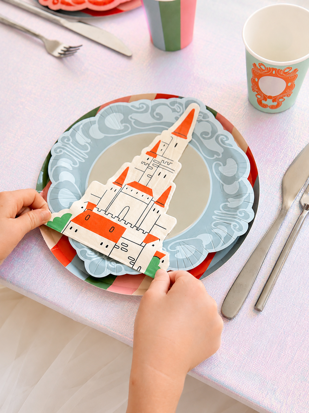Castle shaped paper napkin on stacked blue and red plates with a paper cup and silver cutlery on a pink tablecloth.