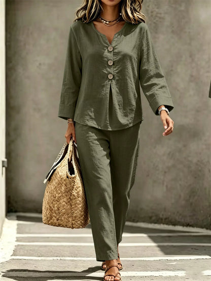 A woman wearing an olive green two-piece linen set with a long-sleeve button-front top and matching wide-leg pants, walking outdoors while carrying a woven straw tote bag.