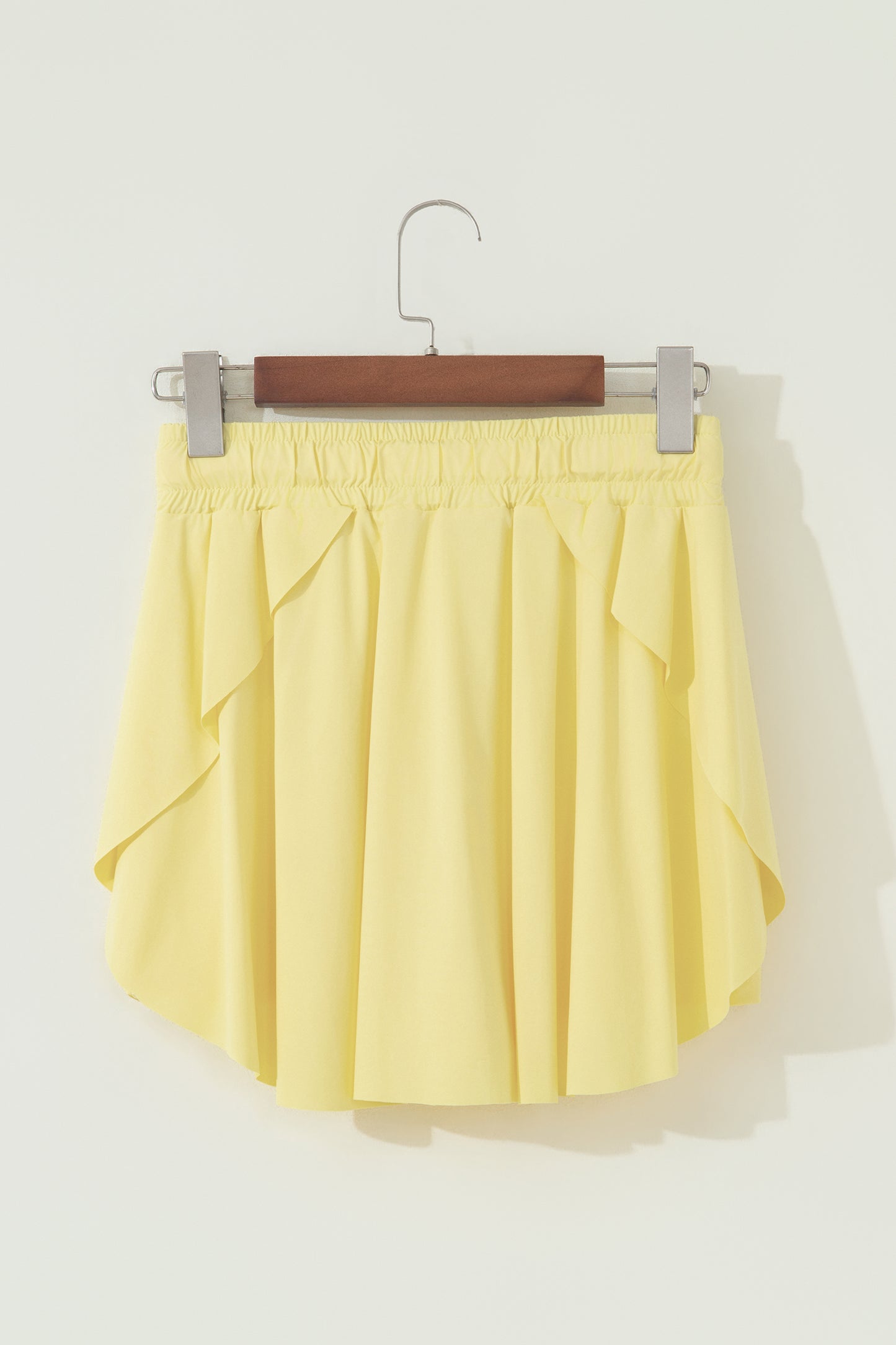 A light yellow pleated mini skirt with side ruffle details, hanging from a wooden hanger with silver clips against a plain off-white wall.