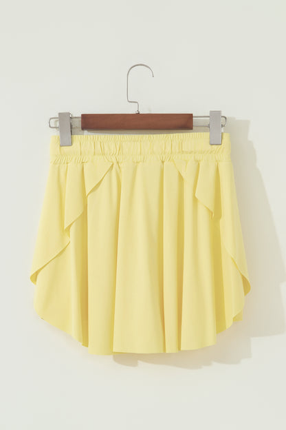 A light yellow pleated mini skirt with side ruffle details, hanging from a wooden hanger with silver clips against a plain off-white wall.
