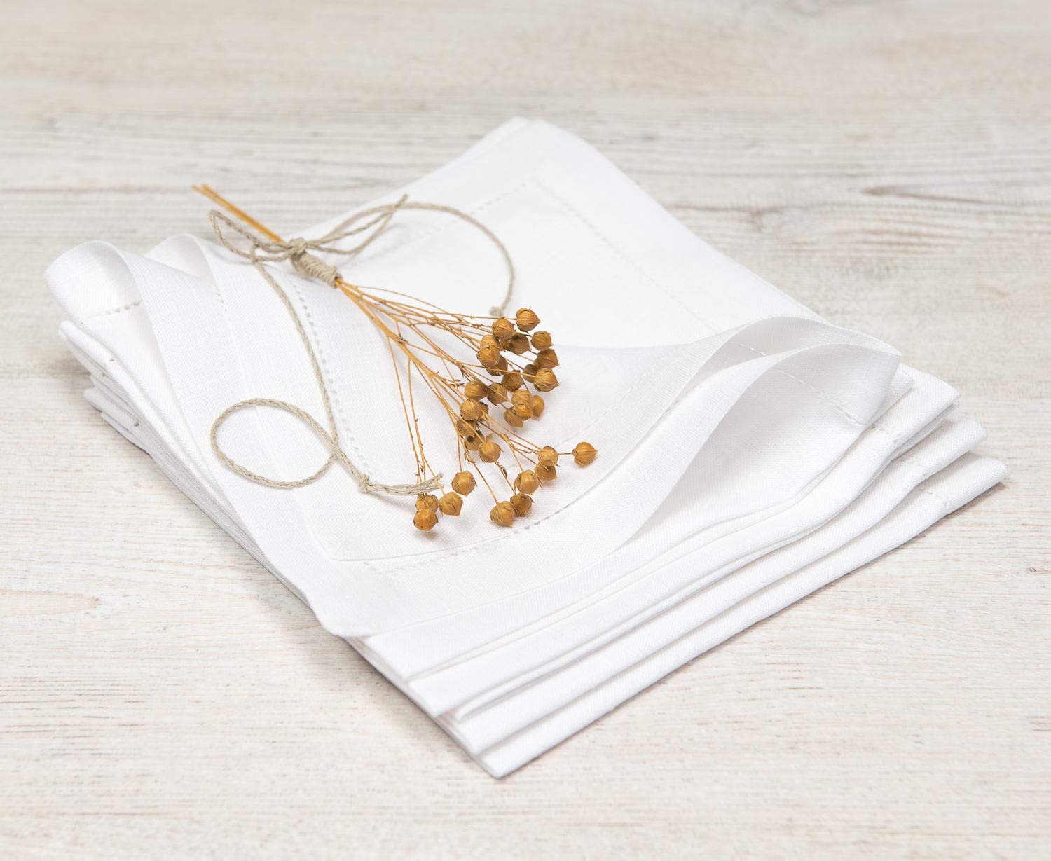 A stack of folded white cotton napkins tied with a thin string and decorated with dried golden-brown flower stems, set on a light wood surface.