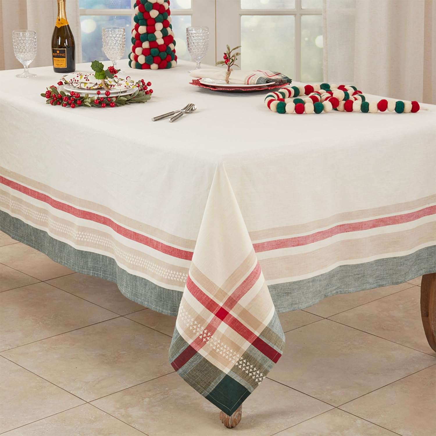 A holiday-themed tablecloth set displayed on a dining table, featuring a white tablecloth with red and beige stripes and a green plaid pattern, along with matching napkins, a festive mini tree, and holiday tableware.