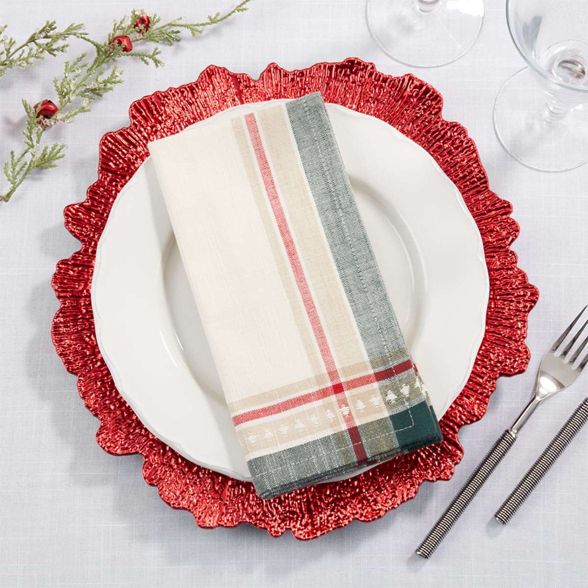 A folded holiday-themed napkin with cream, red, gray, and green plaid and stripe patterns, styled on a white plate over a red scalloped charger plate on a white tablecloth.