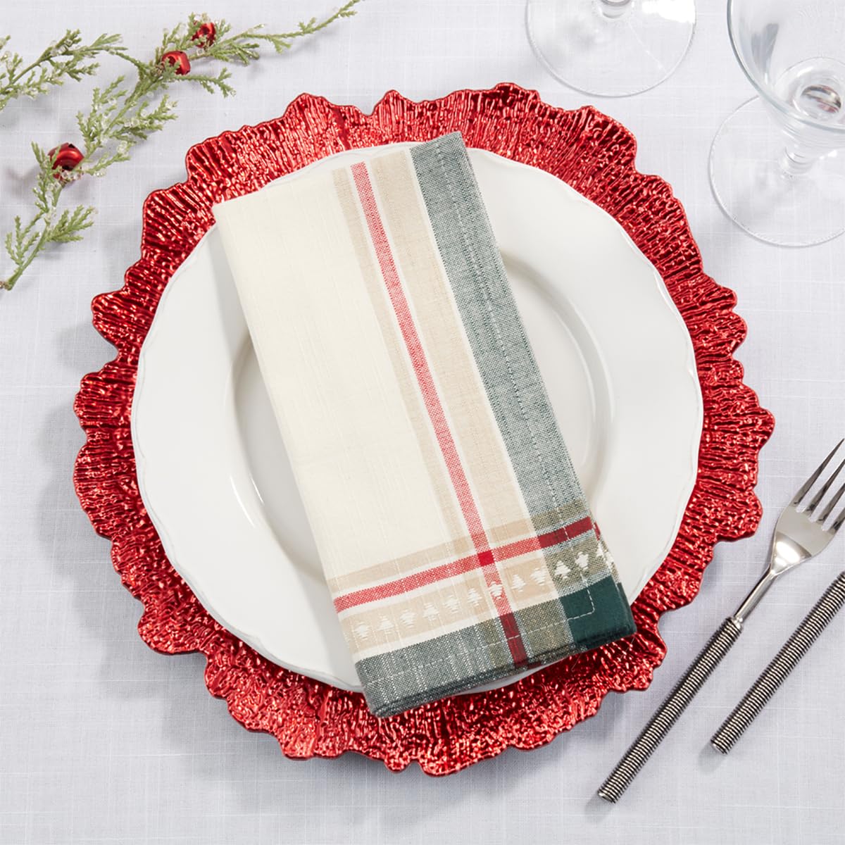A folded holiday-themed napkin with cream, red, gray, and green plaid and stripe patterns, styled on a white plate over a red scalloped charger plate on a white tablecloth.
