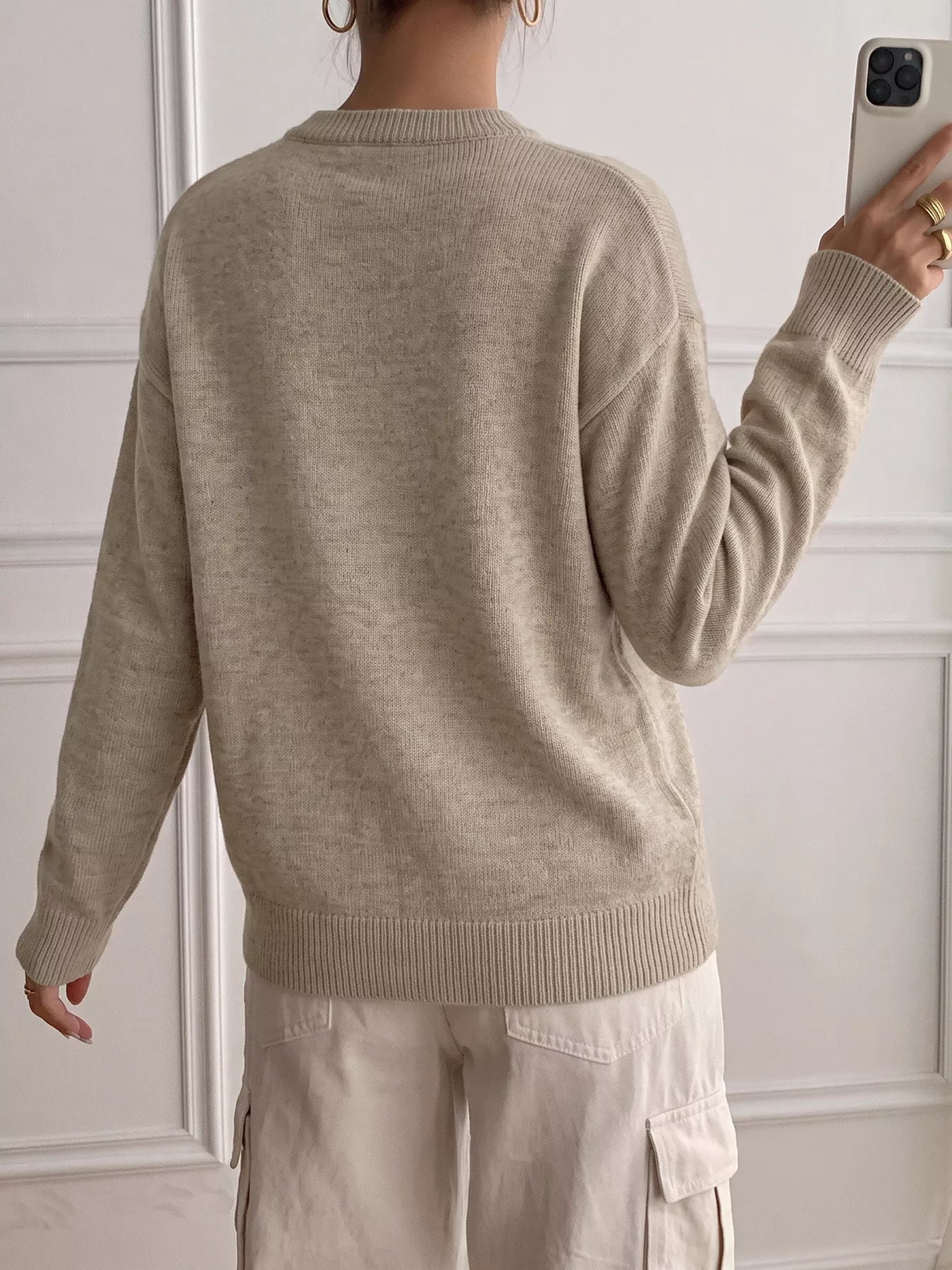 A person shown from the back wearing a light beige knit sweater and matching light cargo pants, standing indoors against a white paneled wall while holding a phone.