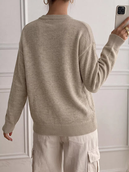 A person shown from the back wearing a light beige knit sweater and matching light cargo pants, standing indoors against a white paneled wall while holding a phone.