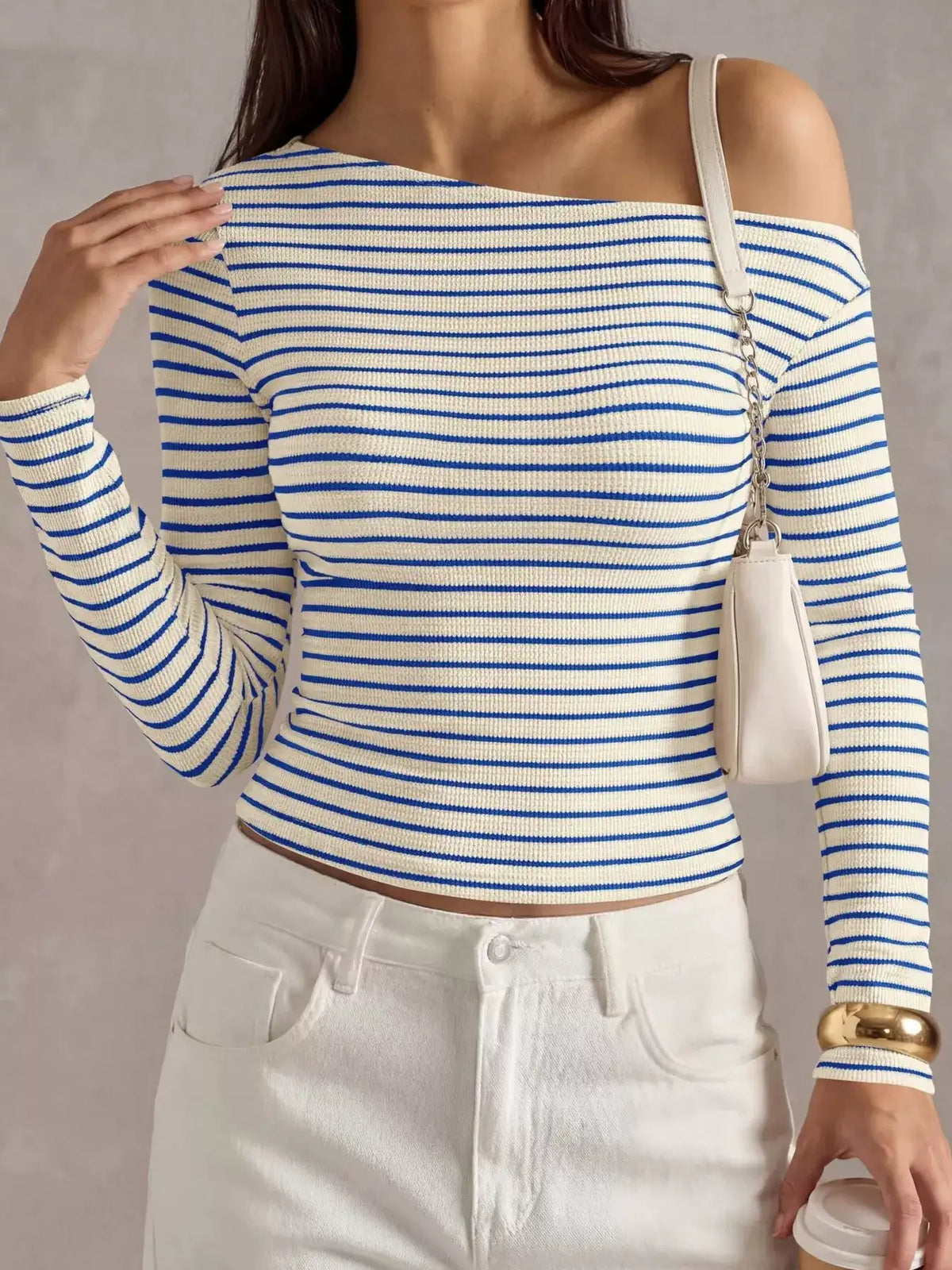 A woman wearing a blue and white horizontally striped off-the-shoulder long-sleeve ribbed top, paired with white pants and a small white shoulder bag.
