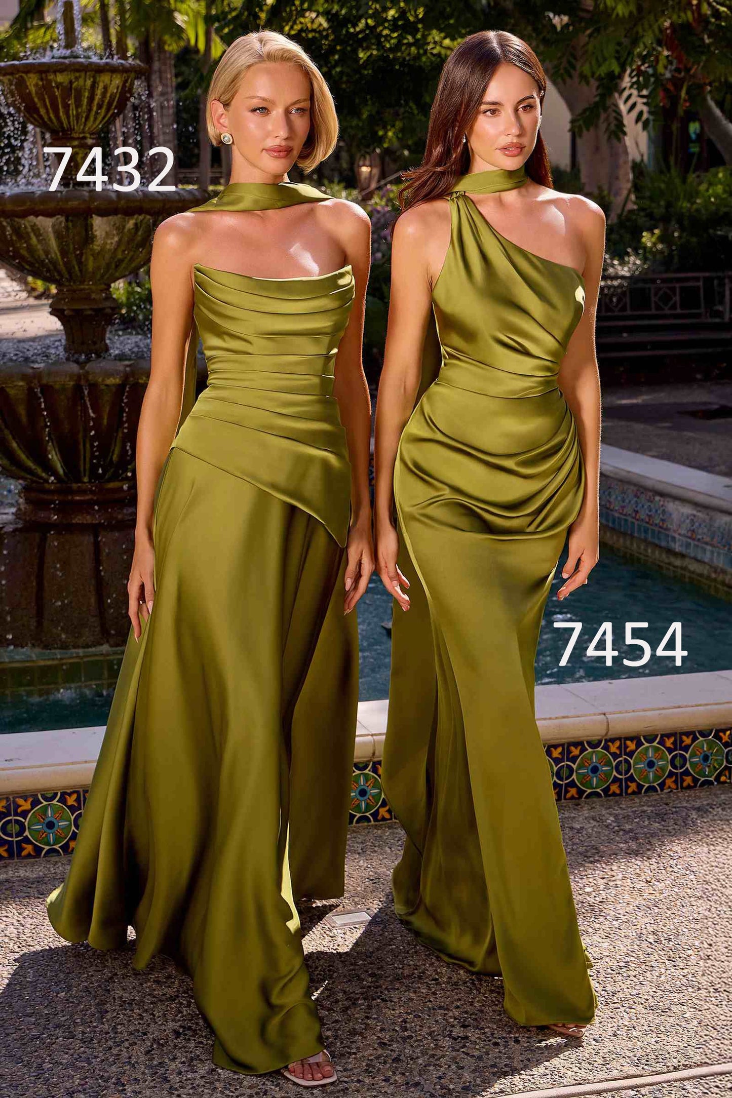 Two women modeling floor-length olive green satin formal dresses in an outdoor courtyard with a fountain and tiled edge visible. One dress is strapless with a draped high neckline and asymmetrical skirt, and the other is a one-shoulder draped gown.