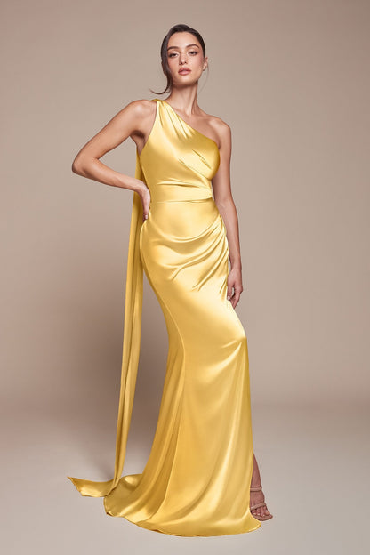 A woman modeling a floor-length yellow satin gown with a one-shoulder draped neckline, gathered waist, and long trailing skirt against a plain beige studio background.