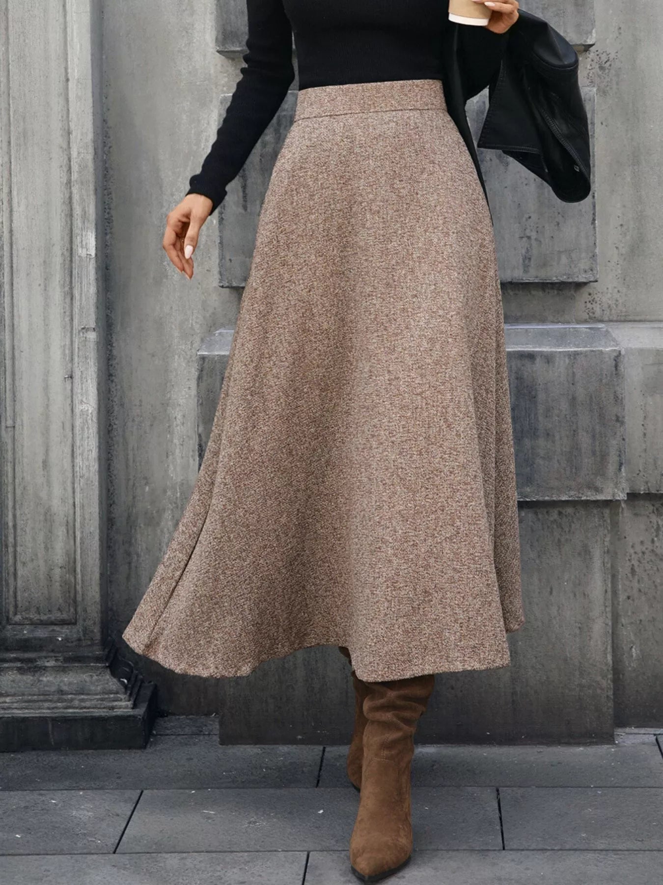 Person wearing a long brown skirt with a black top and brown boots against a gray stone wall.