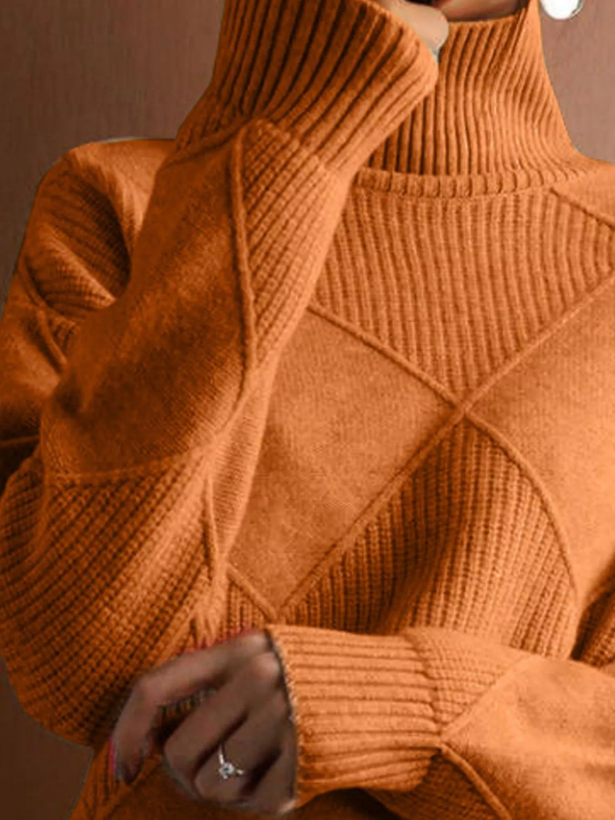 A close-up of a person wearing a chunky knit turtleneck sweater in a warm burnt orange color, featuring diamond-shaped cable knit patterns and ribbed cuffs.