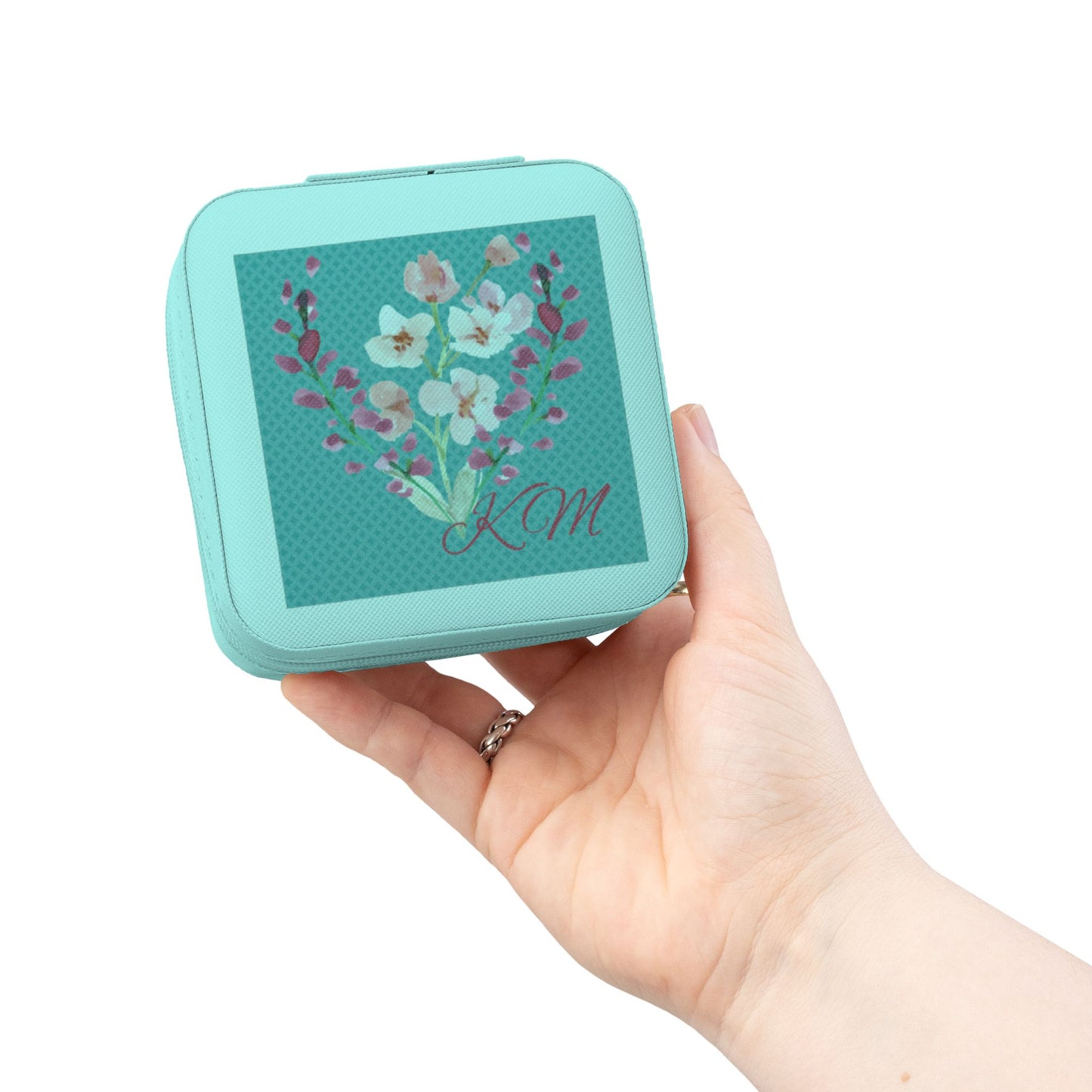 A hand holding a small mint green square case with a floral design and the initials 'KCM' on the front, set against a plain white background.