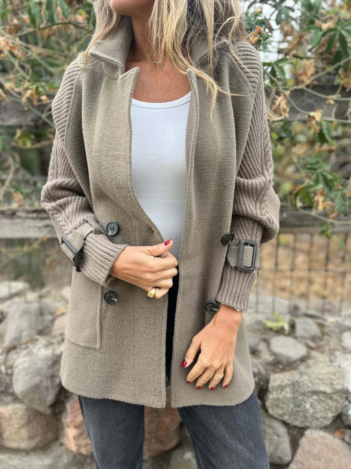 Model wearing a mocha double-breasted coat with cable-knit sleeves, buckled cuffs, wide lapels and front pockets.