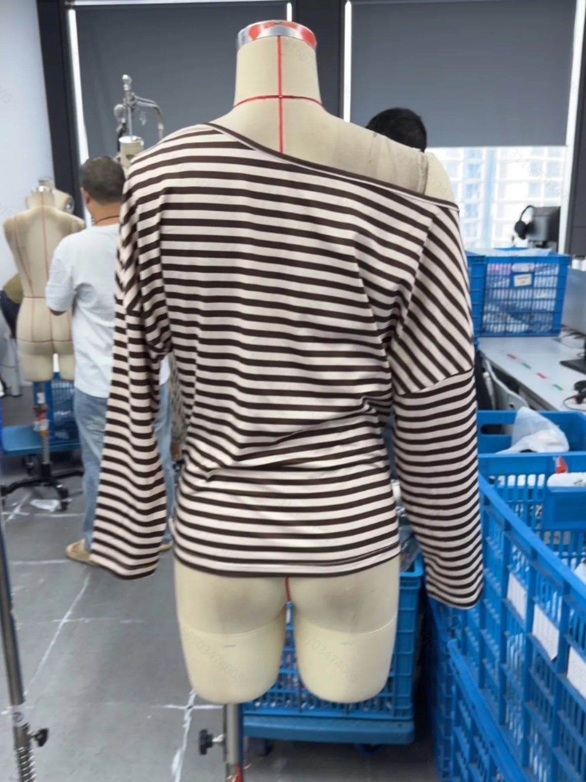 A long-sleeve knit top with horizontal brown and beige stripes, displayed on a dress form mannequin in a tailoring room with other mannequins and blue storage bins visible in the background.