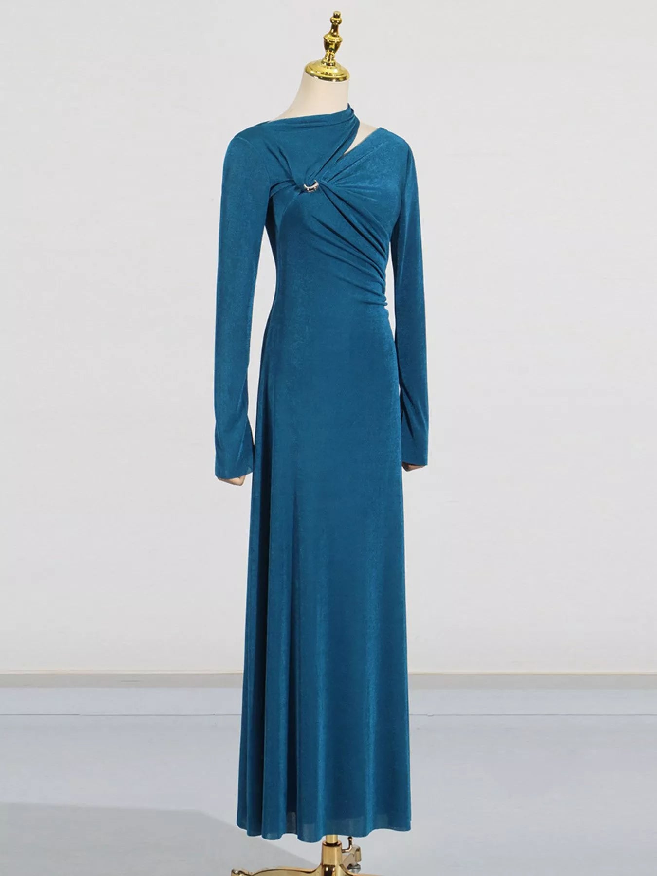 A long-sleeve teal blue maxi dress with a draped crossover front detail and a small silver-toned embellishment, displayed on a mannequin against a plain light gray studio background.