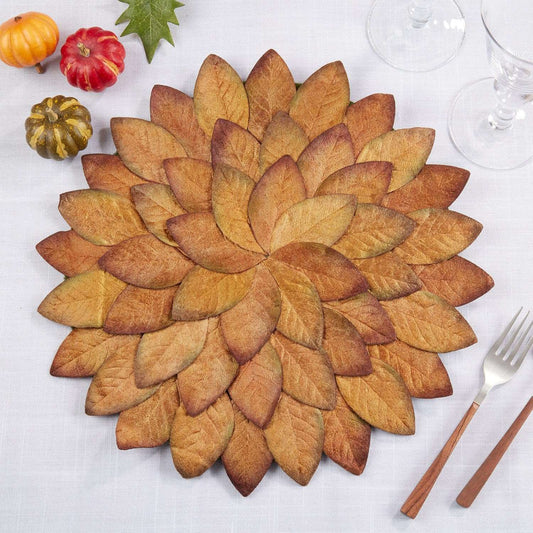 A round autumn-themed placemat made of layered faux leaves in warm fall shades, styled on a white tablecloth with small pumpkins, a green leaf, and silverware.