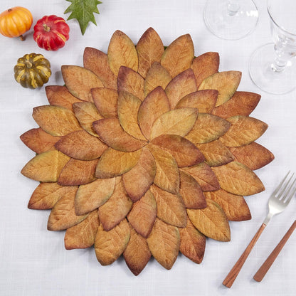 A round autumn-themed placemat made of layered faux leaves in warm fall shades, styled on a white tablecloth with small pumpkins, a green leaf, and silverware.