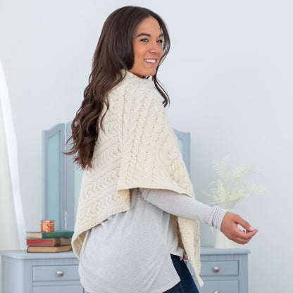 Woman wearing a cream cable knit poncho in a room with a light blue dresser and white walls.