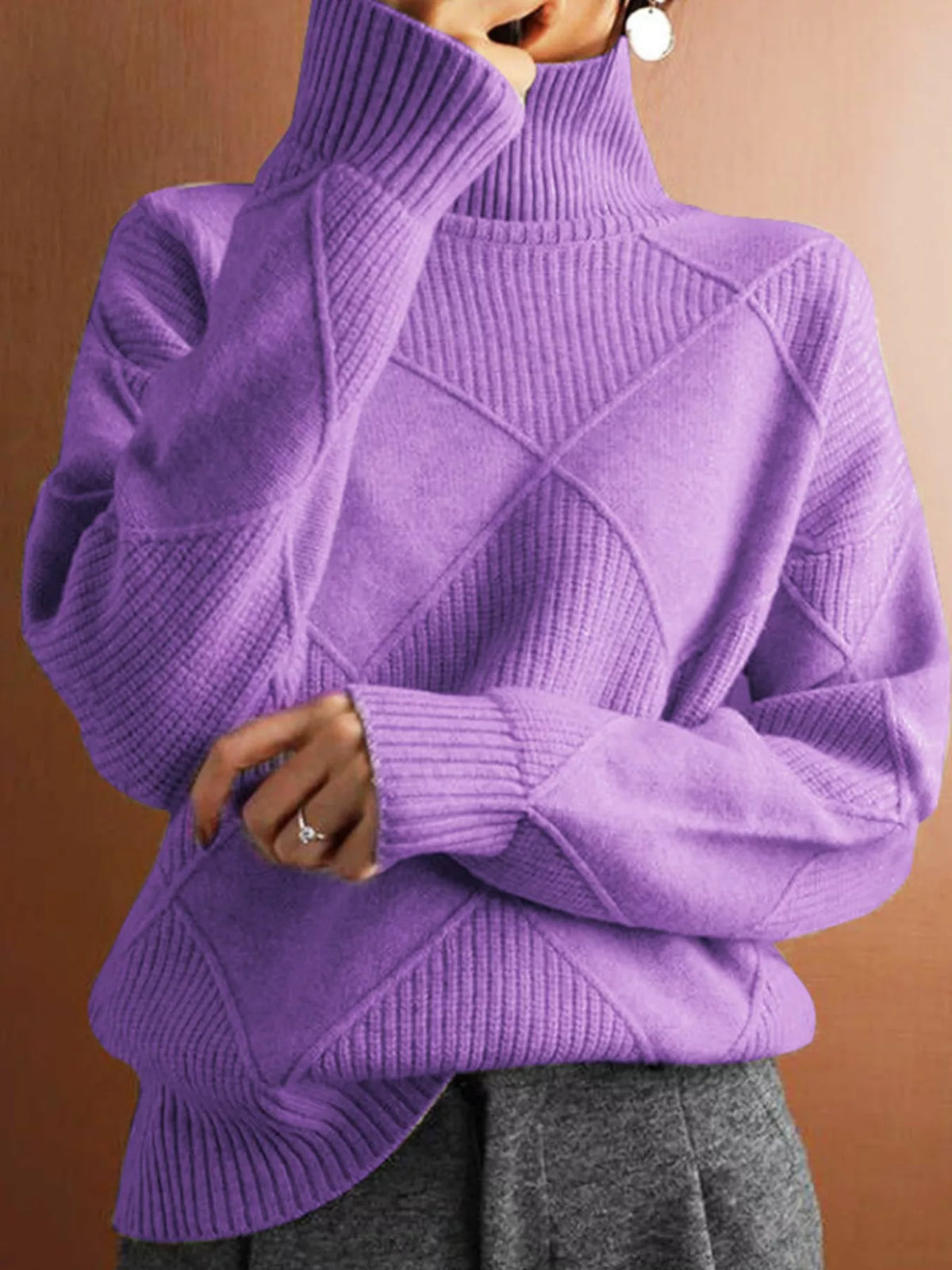 A person wearing a purple ribbed turtleneck sweater with diamond-patterned knit detailing on the front, paired with gray pants, against a warm brown background.