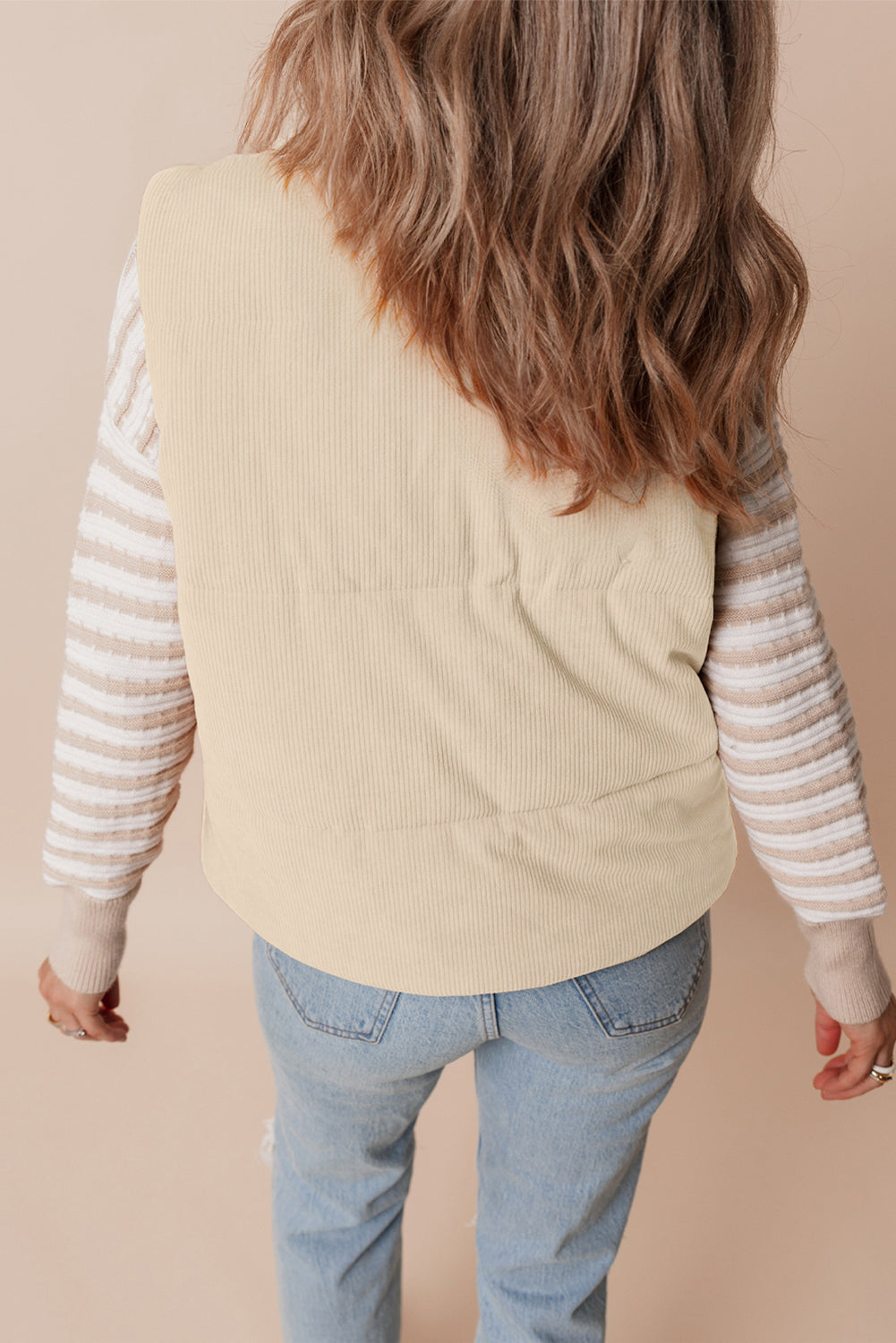 Person wearing a beige vest over a striped shirt with blue jeans on a beige background