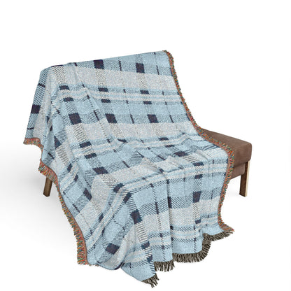 A woven throw blanket with a light blue, gray, and navy plaid pattern and fringed edges, draped over a brown armchair against a white background.