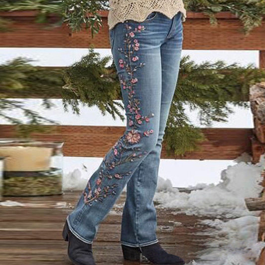 A person wearing blue jeans with pink and green floral embroidery down the front, paired with a cream knit top and black ankle boots, standing on a snowy wooden deck in front of a rustic fence.
