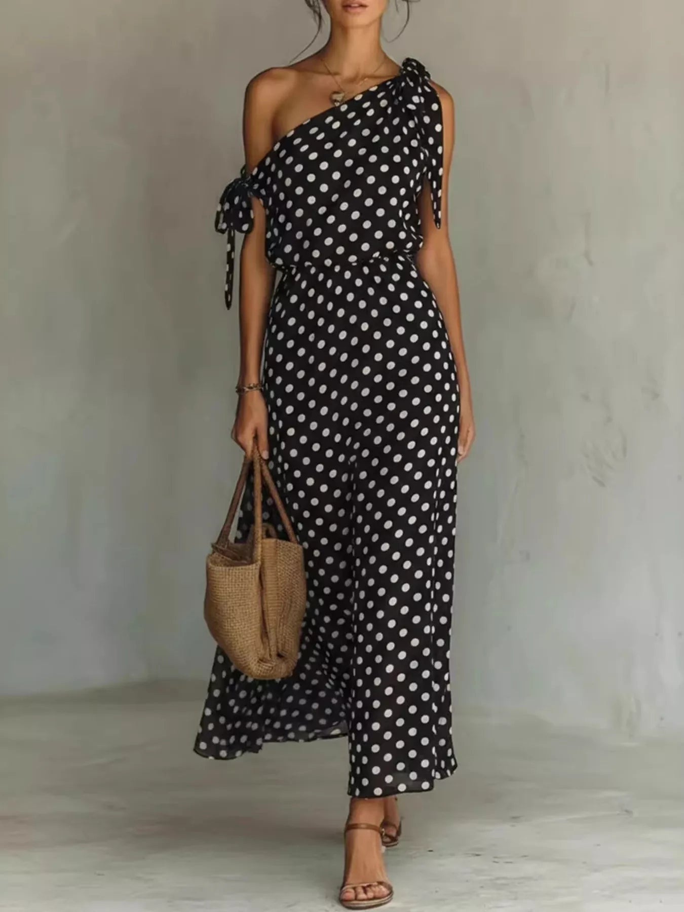 Woman wearing a black polka dot dress holding a woven bag against a plain background
